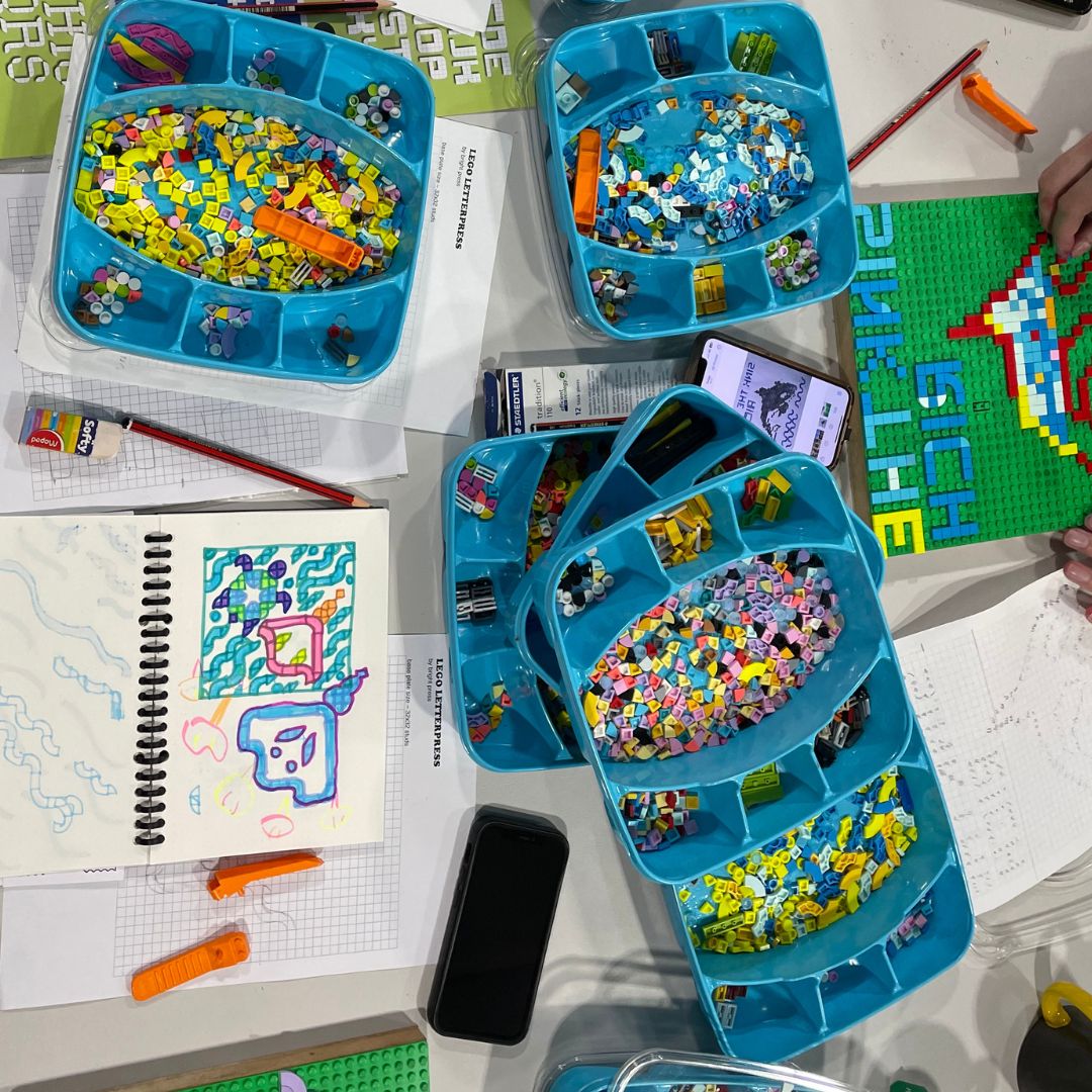 Bird's eye view of a table full of boxes of lego tiles. There is also a notebook of a draft design, and a pair of hands is working on their design on a lego base plate.